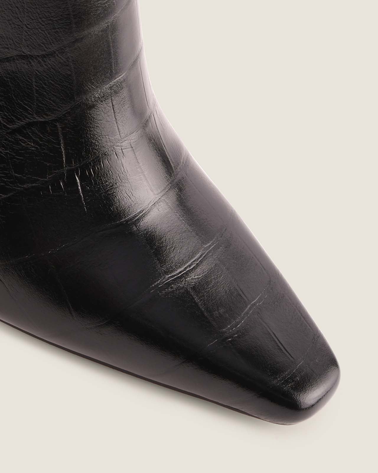 Close-up of a black crocodile leather boot with a pointed toe on a light grey background